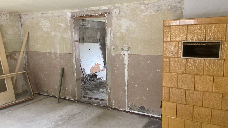 Ein leerer, renovierungsbedürftiger Raum mit einer beschädigten Wand, einer offenen Tür zu einem weiteren unordentlichen Bereich und einem gelblich-braunen, gefliesten Kachelofen auf der rechten Seite. Der Boden besteht aus einfachem Beton, und einzelne Holzrahmenteile sind an der Wand angelehnt.