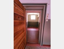 Holztür führt in einen schlichten, kleinen Raum mit rotbraunem Boden, grauen Wänden und einem kleinen Fenster, das Tageslicht hereinlässt; Lichtschalter an der rechten Wand und Zimmertür mit der Nummer 35 beschriftet.