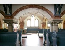 Innenraum einer historischen Kirche mit Ziegelbögen, verzierten Säulen und großen Spitzbogenfenstern. Die hölzernen, blauen Kirchenbänke sind symmetrisch angeordnet, der Boden besteht aus roten Fliesen.