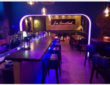 Eine stilvolle Bar mit länglichem Holztresen, umgeben von Barhockern, in einem blau beleuchteten Raum mit violetten Akzenten. Im Hintergrund befinden sich gepolsterte Sitzbänke, Tische und eine leuchtende Wand mit der Aufschrift "La Amistad".