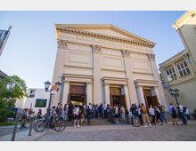 Außenansicht des Maxim Gorki Theaters, einem klassizistischen Gebäude mit Säulenfassade und großen Holztüren, vor denen eine Gruppe von Menschen auf einer breiten Treppe steht. Im Vordergrund sind geparkte Fahrräder und Straßenlaternen zu sehen.