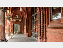 Historische Backsteinarchitektur mit Spitzbögen, verzierten Säulen und einer großen Glastür, die den Eingang zu einem Café markiert; dekorative Elemente wie ein rundes Mosaikfenster und schmuckvolle Kapitelle sind sichtbar.