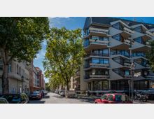 Eine Stadtstraße mit parkenden Autos, flankiert von Bäumen und Gebäuden. Rechts steht ein modernes Wohngebäude mit markanten Balkonen und großflächigen Glasfenstern, links eine Reihe älterer Gebäude mit Bogenfenstern.