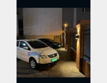 Gepflasterter Parkplatz in einem Hof bei Nacht, beleuchtet durch Wandlampen an Backsteinmauern. Zwei geparkte Autos, ein weißes und ein schwarzes, stehen nebeneinander; im Hintergrund ist eine hohe hellgraue Mauer sowie ein Metalltor zu sehen.