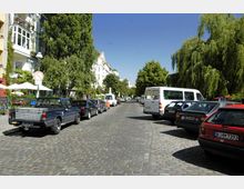 Straßenansicht mit Kopfsteinpflaster, parkenden Autos auf beiden Seiten und mehrstöckigen Wohnhäusern mit Balkonen und Begrünung auf der linken Seite, sowie großen Bäumen entlang der Straße. Ein blauer Himmel und Sonnenschein dominieren die Szene.
