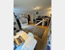 Ein Kinderzimmer mit Holzfußboden, einem grauen Teppich in der Mitte und einem Hochbett rechts. Links befindet sich ein Schreibtisch mit Laptop, darüber hängen gerahmte Bilder an der Wand, während Regale und Aufbewahrungsmöbel das Zimmer strukturieren.