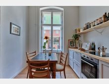 Schmale Küche mit großem weißen Fenster, das auf ein Gebäude mit grüner und roter Fassade blickt. Der Raum enthält einen Holztisch mit vier Stühlen, offene Regale mit Gewürzgläsern und eine weiße Küchenzeile mit Herd und Backofen; eine Sonnenblume steht in einer Vase auf dem Tisch.