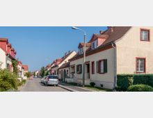 Eine ruhige Wohnstraße mit Reihenhäusern im traditionellen Stil, hellen Fassaden, grünen Fensterläden und roten Dächern. Entlang der Straße sind geparkte Autos, Laternen und vereinzelte Begrünung zu sehen.