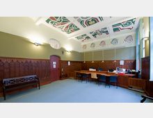 Ein Büroraum mit hohen Decken, verziertem Deckenmuster und Holzvertäfelung an den Wänden. Der Raum enthält einen großen Holztisch mit mehreren Stühlen und Büromaterialien sowie eine Holzbank an der Wand.