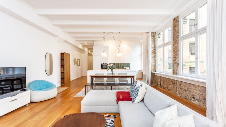 Heller, offener Loft-Wohnraum mit Küche und Essbereich, weißen Deckenbalken und Holzboden. Große Fensterfront mit freigelegter Ziegelwand sowie moderne Einrichtung mit Sofa, Couchtisch und Kücheninsel.
