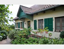 Einfamilienhaus mit cremefarbener Fassade, dunkelgrünen Fensterläden und Ziegeldach, umgeben von einem gepflegten Garten mit Büschen, Blumen und Zierpflanzen. Ein kleiner gepflasterter Weg führt durch den Garten zur Eingangstür.