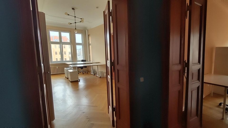 Blick durch einen Flur in ein helles Büro mit Fischgrät-Holzboden, hohen Decken und großen Sprossenfenstern. Im Raum stehen mehrere Schreibtische und Bürostühle; rechts sind geöffnete dunkle Holztüren und ein weiterer Arbeitsbereich zu sehen.