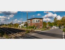 Ziegelgebäude eines Stellwerks mit der Aufschrift "Zossen" an einem Bahnübergang, flankiert von Bahngleisen mit Oberleitungen und einer Schranke. Im Hintergrund sind Bäume, vereinzelte Gebäude und ein bewölkter Himmel zu sehen.