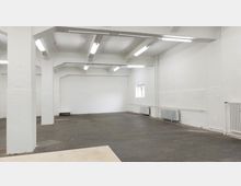 Ein leerer, rechteckiger Raum mit weißen Wänden, sichtbaren Heizkörpern entlang der rechten Wand und einer dunkelgrauen gefliesten Bodenfläche. Die Decke wird von weißen Trägern gestützt und ist mit länglichen Leuchtstofflampen beleuchtet.