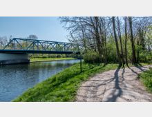 Flusslandschaft mit einer blauen Stahlbrücke im Hintergrund, die den Fluss überspannt. Am Ufer sind ein grüner Rasen, ein Schotterweg und mehrere hohe, blattlose Bäume zu sehen, die von Sonnenlicht durchbrochen werden.