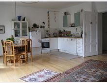 Offene Wohnküche mit Holzfußboden, einem Esstisch aus Holz mit vier Stühlen, einem Teppich mit buntem Muster und einer Küchenzeile mit weißen Schränken, einem Kühlschrank, einem Herd und einer Spülmaschine. Die Wände sind weiß, und es stehen dekorative Gegenstände auf den Möbeln.