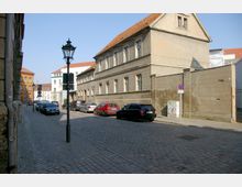 Straßenszene mit gepflastertem Gehweg und Straße, gesäumt von historischen Gebäuden mit roten Ziegeldächern und schlichten Fassaden. Autos sind am Straßenrand geparkt, und eine klassische Straßenlaterne steht in der Mitte des Gehwegs.