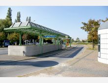 Haltestelle "U-Bhf Innsbrucker Platz" mit grünem Stahl- und Glasüberdachungssystem, umgeben von Bäumen und einem gepflasterten Gehweg; im Hintergrund eine Straße und ein offenes Areal.