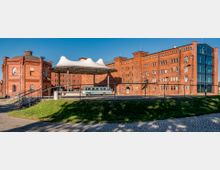 Ziegelbaukomplex mit historischen Gebäuden und modernen Elementen; links ein Rundbau mit Kuppel, rechts ein großes Backsteingebäude mit Glasfassade an der Seite. In der Mitte eine moderne Überdachung mit einem nostalgischen Bus darunter, umgeben von Kopfsteinpflaster und Rasen.