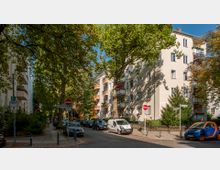 Straßenansicht mit mehrstöckigen Wohngebäuden aus hellem Putz, Bäumen mit dichtem Laub und parkenden Autos an beiden Seiten der Straße. Verkehrsschilder und ein Gehweg ergänzen den urbanen Charakter der Szene.