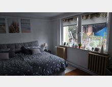 Schlafzimmer mit großem Bett in grauer Bettwäsche, weißen Wänden und drei Wandbildern über dem Kopfteil. Rechts eine breite Fensterfront mit Fensterbank voller kleiner Dekorationen und Blick in einen Garten; darunter stehen zwei Heizkörper.