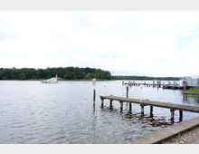 Ein ruhiger See mit einem bewaldeten Ufer im Hintergrund, einem weißen Motorboot auf dem Wasser und mehreren hölzernen Stegen am rechten Bildrand. Der Himmel ist bewölkt, und der Steg im Vordergrund führt ins Wasser.