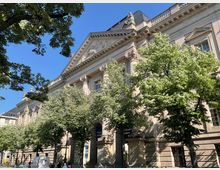 Historisches Gebäude mit klassizistischer Fassade, verziert durch Säulen, ein Giebeldreieck und dekorative Reliefs, umgeben von Bäumen an einer Straße bei klarem Himmel.