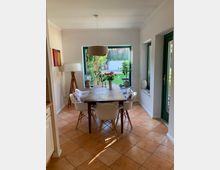 Essbereich mit einem Holztisch, umgeben von vier weißen Stühlen, einer Stehlampe und einer Hängelampe. Große Fenster mit grünem Rahmen bieten Blick auf einen Garten mit Pflanzen und einer kleinen Terrasse; der Boden ist mit hellbraunen Fliesen versehen.