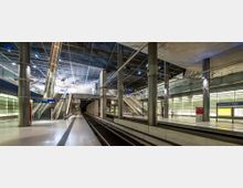 Moderner U-Bahnhof mit breiten Bahnsteigen, gläsernen Wänden, metallischen Säulen und einer Rolltreppe, die zu einer höheren Ebene führt. Eine Beschilderung zeigt „Potsdamer Platz“, und der Bereich ist von künstlichem Licht erhellt.