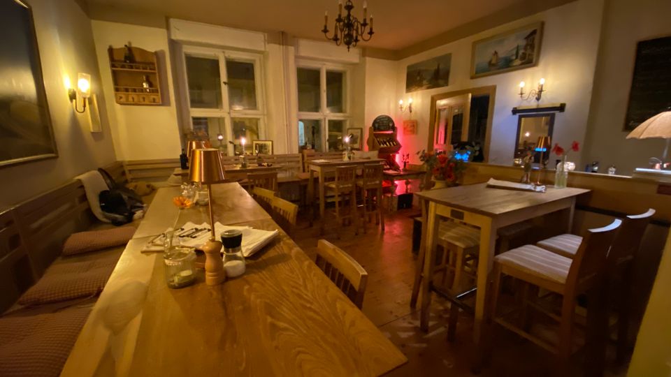 Gemütlicher Gastraum eines Restaurants mit mehreren Holztischen, Sitzbank und Holzstühlen auf Holzboden. Warmes Licht von Wandleuchten und einem Kronleuchter, dazu große Fenster, gerahmte Bilder an den Wänden und ein Spiegel.
