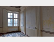 Ein leerer, verfallener Raum mit hohen Wänden, verzierten Pfeilern und einem großen rechteckigen Sprossenfenster. Die Wände sind beschädigt und mit Graffiti besprüht, der Boden zeigt abblätternde Farben.