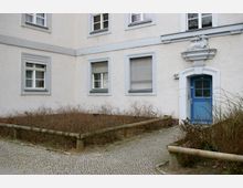 Wohnhausfassade mit heller Putzfassade, mehreren Fenstern mit teilweise geschlossenen Rollläden, und einem blauen Eingangstürportal. Über der Tür befindet sich ein kleiner steinerner Schmuckaufsatz mit einem Relief aus zwei Tieren, umgeben von einem bepflanzten Vorgarten mit niedriger Holzabsperrung.