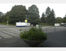 Ein offener Platz mit gepflastertem Boden, im Vordergrund ein großer, runder Pflanzkübel mit grünem Bewuchs. Im Hintergrund befinden sich Bäume, eine Straße mit parkenden Anhängern und ein geparkter Lieferwagen.