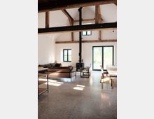 Wohnraum mit offenem Dachstuhl aus Holz, einem freistehenden schwarzen Kaminofen und großen Fenstern, die viel Tageslicht hereinlassen. Der Raum ist modern eingerichtet mit einer braunen Couch, einem Esstisch aus Holz und minimalistischem Mobiliar auf einem polierten Betonboden.