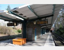 U-Bahn-Station mit überdachtem Bahnsteig, sichtbaren Anzeigetafeln für die Linie U3 und einer Uhr; im Hintergrund Gleise und eine grüne, bewaldete Umgebung. Die Architektur umfasst Ziegelwände und metallene Träger unter einem hellen Wellblechdach.