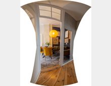 Blick durch eine hohe, weiß gerahmte Glastür in einen Wohn‑ und Essbereich mit gemusterten Fliesen, dunkler Akzentwand und gerahmten Bildern. Im Raum hängen eine runde Pendelleuchte, davor stehen ein Esstisch mit gelben Stühlen und daneben ein Sofa; im Vordergrund liegt ein Dielenboden.