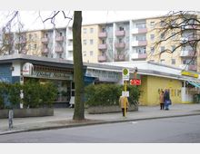 Straßenszene mit kleinen Geschäften, darunter das „Pichel Stübchen“ und eine Eisdiele namens „Casa del Gelato“. Im Hintergrund ein mehrstöckiges Wohngebäude mit Balkonen, im Vordergrund Gehweg, Bäume und eine Bushaltestelle.
