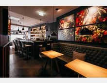 Modernes Bar- und Restaurantinterieur mit langem Tresen, Barhockern und beleuchteten Flaschenregalen. Rechts stehen Holztische vor einer gepolsterten Sitzbank, darüber hängen schwarze Pendelleuchten und großformatige Wandbilder mit Pflanzen- und Gewürzmotiven.