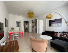 Wohnzimmer mit Parkettboden, einer schwarzen Couch, einem ovalen weißen Couchtisch und einem rosa Sessel. An der Decke hängt eine auffällige goldene Lampe, im Hintergrund steht ein roter Esstisch mit weißen Stühlen vor einem Bücherregal. Türen und Wände sind weiß gestrichen.