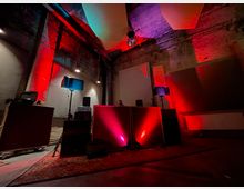 Ein industrieller Innenraum mit unverputzten Backsteinwänden, rot-violetter Beleuchtung und Tonstudio-Equipment, darunter Lautsprecher und Mischpulte, auf einem Teppichboden. Die Atmosphäre deutet auf einen Veranstaltungs- oder Musikraum hin.