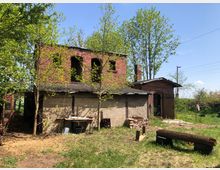 Backstein- und Lehmgebäude auf einem grasbewachsenen Grundstück, umgeben von jungen Bäumen. Im Vordergrund befinden sich Holzbänke, ein Tisch sowie gestapelte Ziegel, während im Hintergrund Stromleitungen und ein klarer Himmel zu sehen sind.