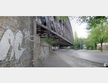 Eine stählerne Eisenbahnbrücke mit Graffiti überspannt eine gepflasterte Straße, die von Bäumen gesäumt ist. Links im Bild befindet sich eine mit Graffiti versehene gemauerte Wand.