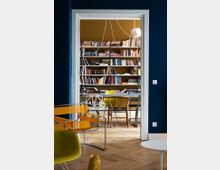 View through a white doorway into a home library or study with built-in wall-to-wall bookshelves, a central table with mixed modern chairs, and a hanging pendant light. The room has a herringbone wood floor and dark blue walls with colorful seating visible in the foreground.