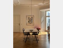 Helles Esszimmer mit Holzboden, vier modernen Freischwinger-Stühlen um einen weißen Tisch und einer Hängelampe darüber. Ein großes Fenster und eine Glastür lassen Tageslicht herein; an der Wand hängt ein gerahmtes Kunstwerk.