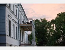 Seitenansicht eines historischen Gebäudes mit weißer Putzfassade, verzierten Fensterrahmen und zwei Balkonen mit Metallgeländern. Im Hintergrund sind große grüne Bäume und ein Himmel bei Sonnenuntergang zu sehen.