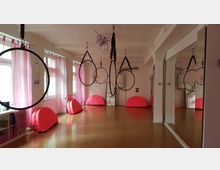 Ein heller Trainingsraum mit Holzboden und großen Spiegeln an der Wand. Mehrere Gymnastikringe sind von der Decke abgehängt, und entlang der Fenster mit rosa Vorhängen stehen rote gepolsterte Matten.