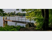 Ein kleiner Metallsteg mit Geländer ragt von einem bewaldeten Ufer in einen ruhigen See. Im Hintergrund sind Bäume, ein Strand und ein Boot auf dem Wasser zu sehen.
