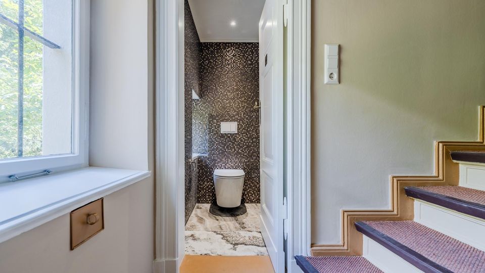Blick vom Treppenhausflur in ein kleines, modernes Gäste-WC mit dunkler Mosaikfliesenwand, wandhängendem WC und hellem Steinboden. Links befindet sich ein großes Fenster, rechts eine Treppe mit Teppichläufer und Holzstufenkanten.