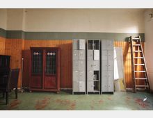 Ein rustikaler Raum mit holzvertäfelten Wänden, grüner abgenutzter Bodenfarbe und einer Reihe von grauen Metallschränken. Neben den Schränken stehen ein Holzschrank mit verzierten Glastüren, eine Leiter und ein schwarzes Klavier links im Bild.