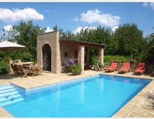 Privater Gartenbereich mit einem rechteckigen Pool, umgeben von einer Terrasse aus beigen Steinfliesen. Im Hintergrund stehen eine gemauerte Backsteinpergola mit Holzdach, mehrere Liegen mit roten Polstern und ein Holztisch mit Klappstühlen unter einem Sonnenschirm; umgeben von grünen Bäumen und Blumen.